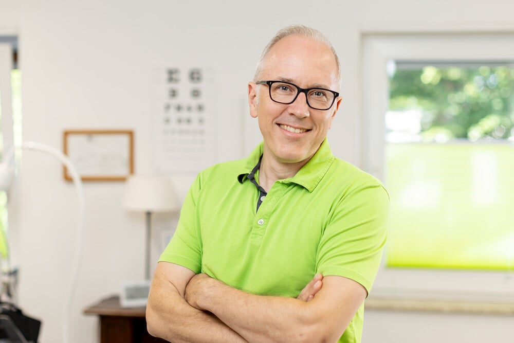 Ein Mann in einem hellgrünen Polohemd steht mit verschränkten Armen in einem Raum mit medizinischen Geräten und einer Sehtesttafel im Hintergrund