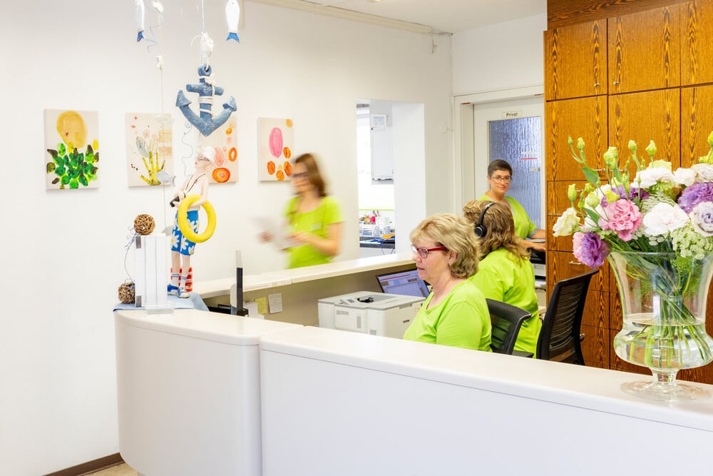 Ein heller Empfangsbereich mit Mitarbeitern in hellgrünen Shirts, dekorativen Gemälden und einem großen Blumenarrangement auf der Theke