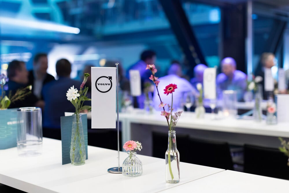 Ein modernes Event-Setup mit einem weißen Tisch, der mit Blumenarrangements und einer Volvo-Flagge geschmückt ist und verschwommene Menschen im Hintergrund