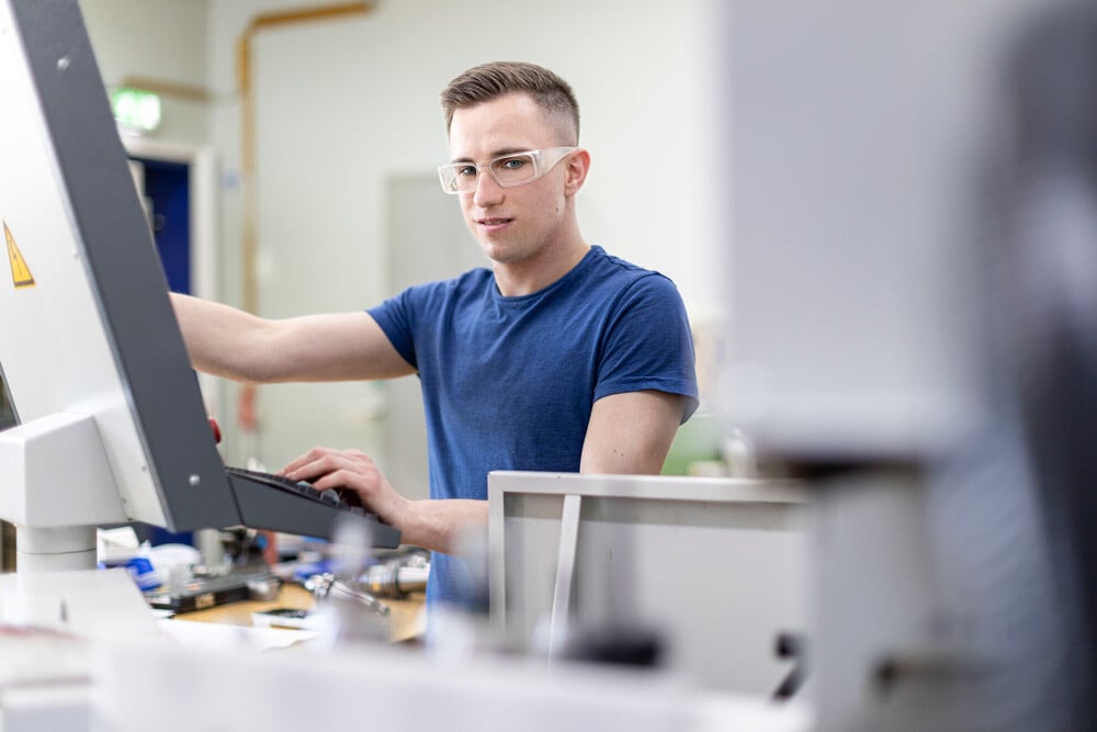 Eine Person in einem blauen Hemd bedient einen Computer in einer Werkstatt voller Werkzeuge und Maschinen mit Blick in die Kamera